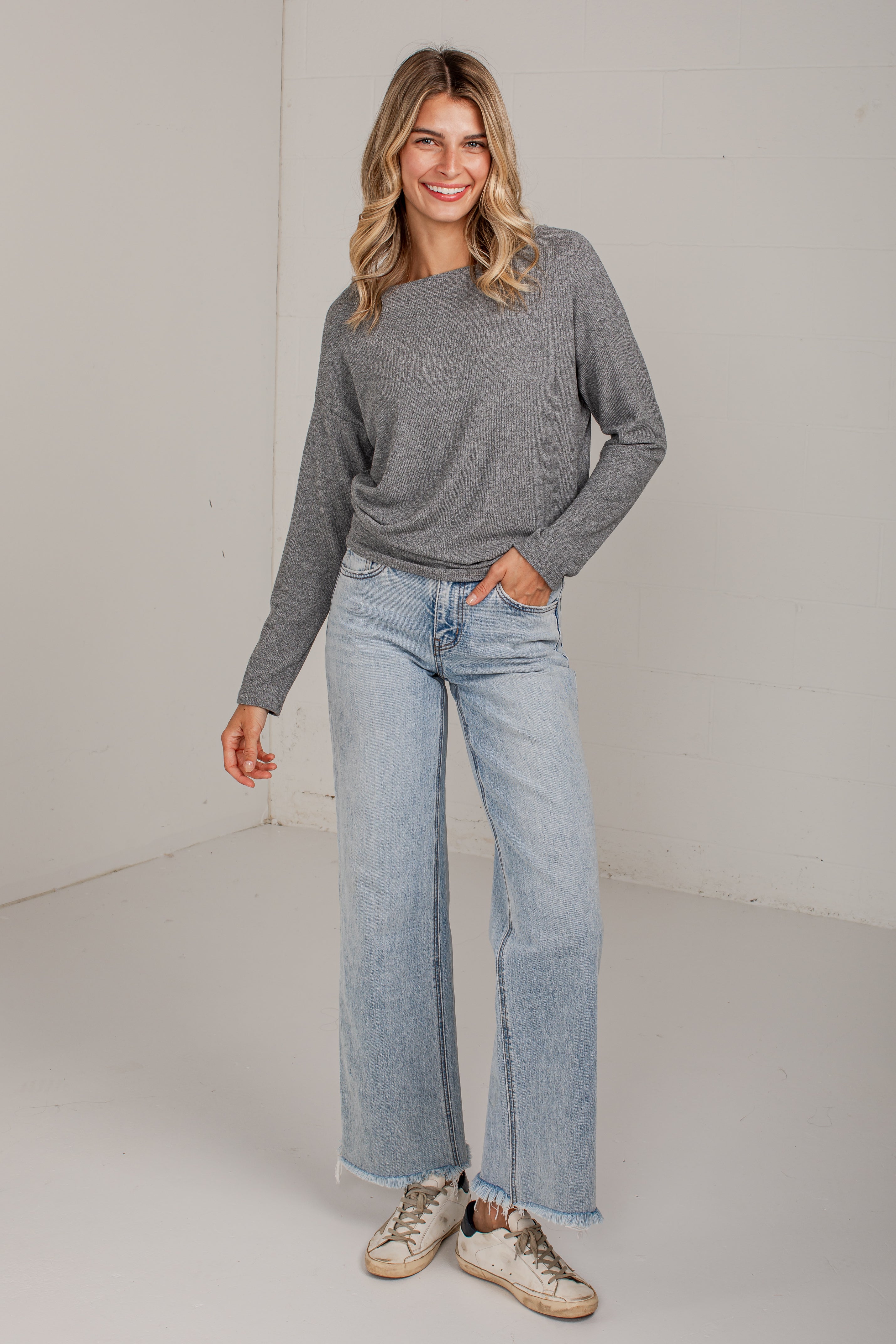 A woman modeling a gray long-sleeve knit top and light-wash wide-leg jeans with beige sneakers, standing against a plain off-white studio wall.