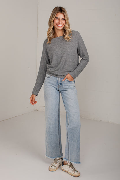 A woman modeling a gray long-sleeve knit top and light-wash wide-leg jeans with beige sneakers, standing against a plain off-white studio wall.
