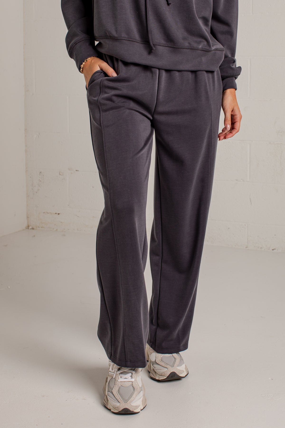 A person wearing dark charcoal gray sweatpants with a matching sweatshirt and white sneakers, standing in a minimalist studio setting with a white brick wall background.