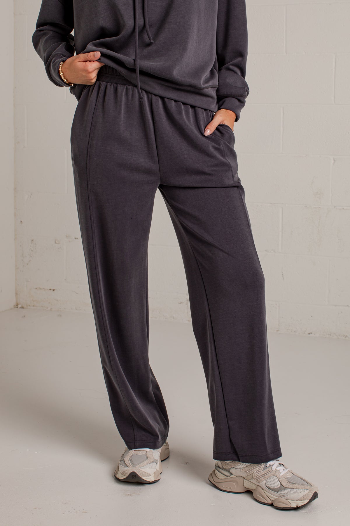 A person wearing dark charcoal gray wide-leg sweatpants with a matching sweatshirt and chunky off-white sneakers, standing against a light gray brick wall.