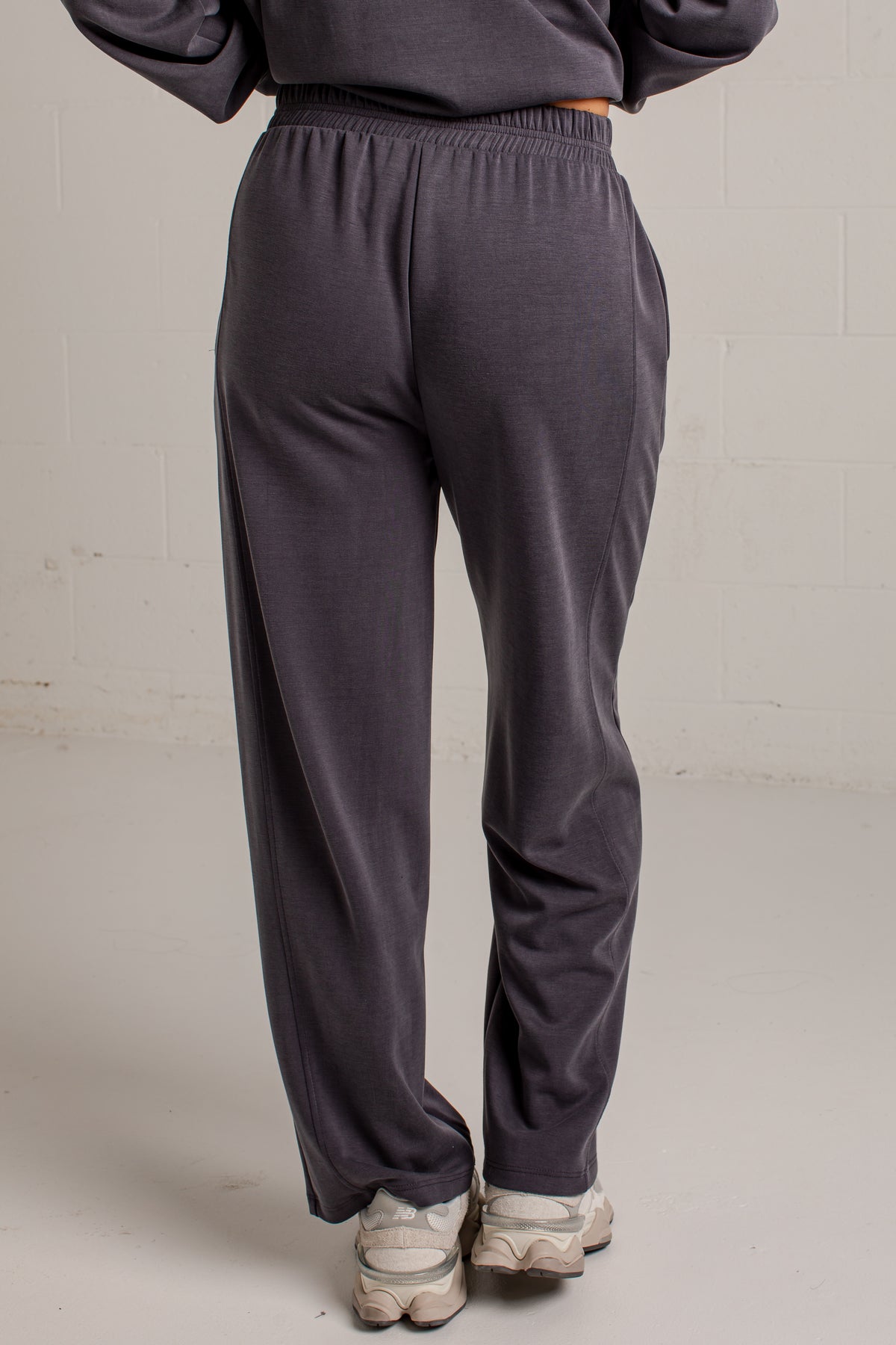 A person shown from the back wearing dark charcoal gray sweatpants and a matching sweatshirt, standing against a light gray wall.