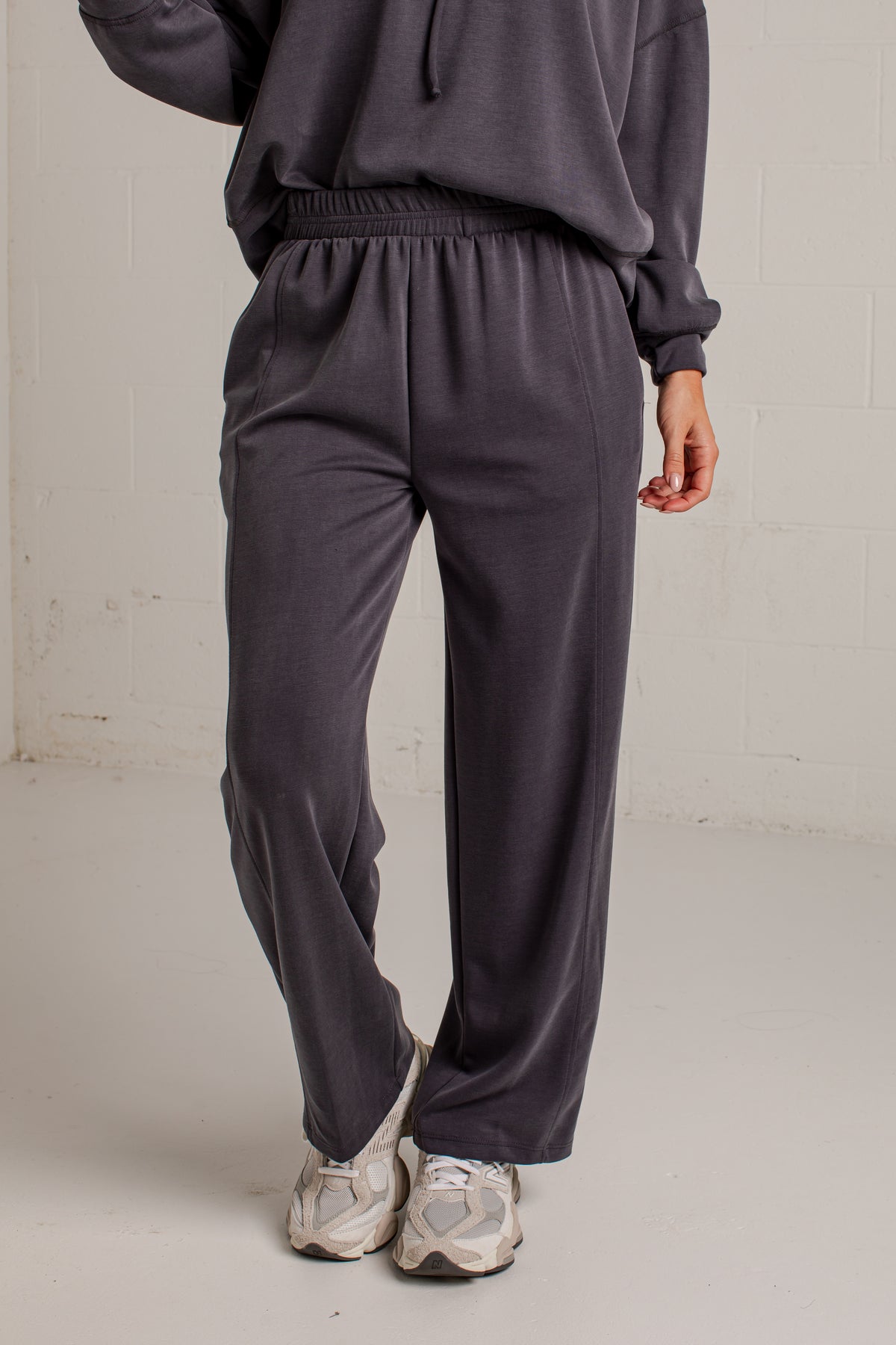 A person wearing dark charcoal gray sweatpants and a matching sweatshirt, standing in a studio with a light gray floor and white brick wall background.
