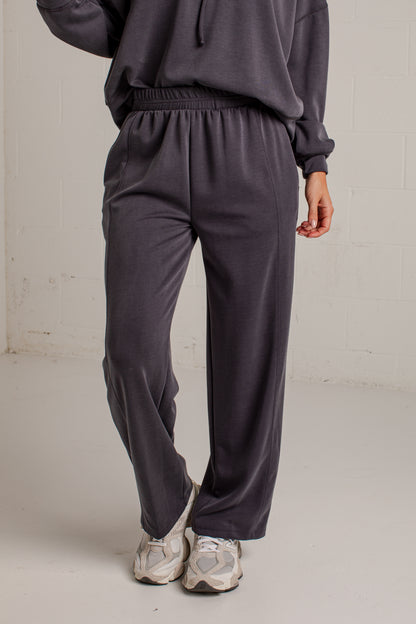 A person wearing dark charcoal gray sweatpants and a matching sweatshirt, standing in a studio with a light gray floor and white brick wall background.