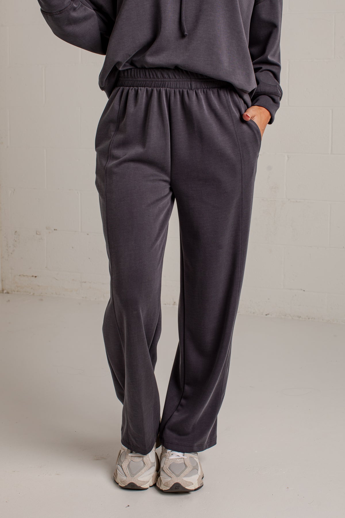 A person wearing dark charcoal gray sweatpants with an elastic waistband and side pockets, paired with a matching dark hoodie and light gray sneakers, standing against a light gray brick wall.