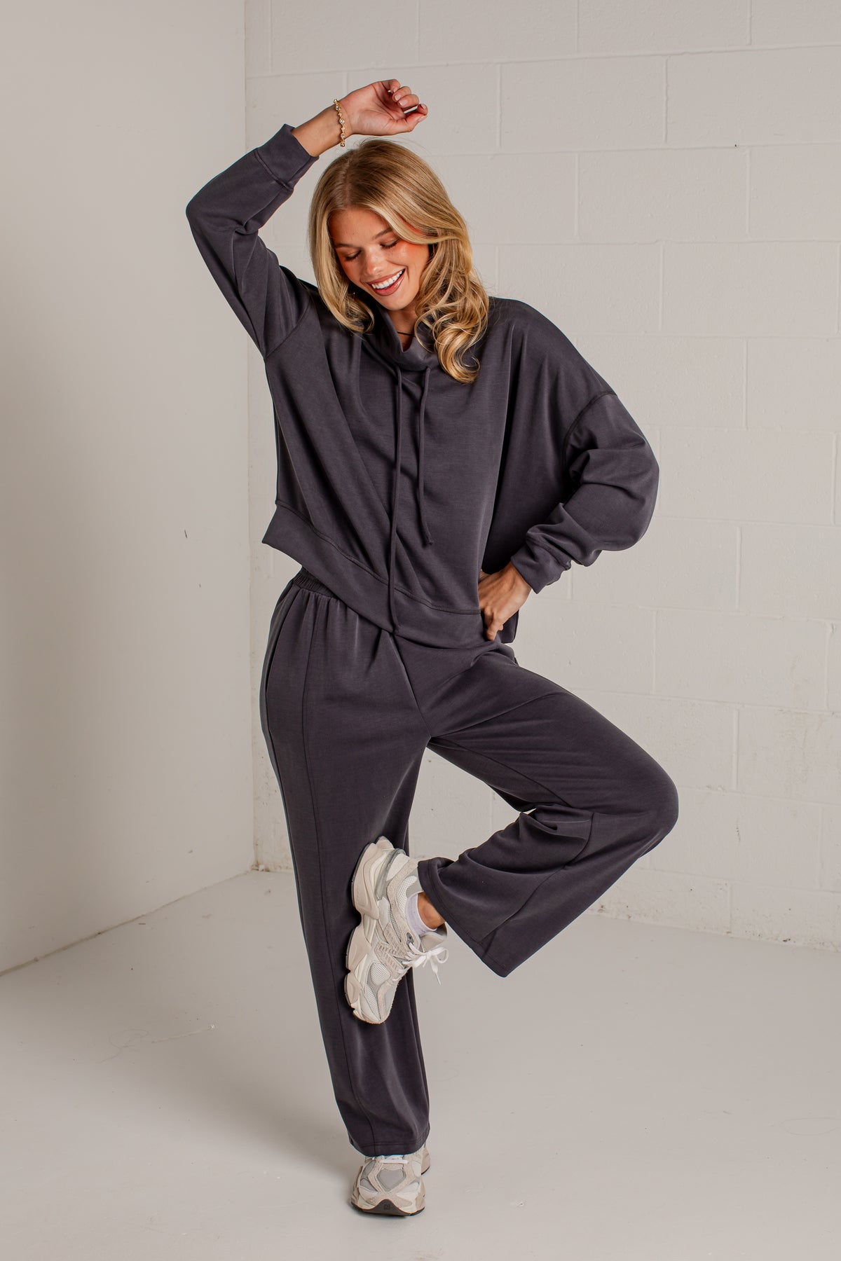 A woman modeling a dark gray two-piece loungewear set consisting of a cropped hoodie and matching sweatpants, paired with white sneakers, in a bright studio setting.