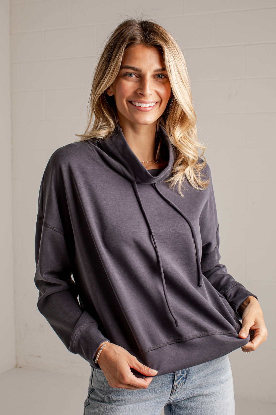 A woman wearing a dark charcoal gray oversized pullover hoodie with drawstrings, paired with light blue jeans, standing against a white brick wall.