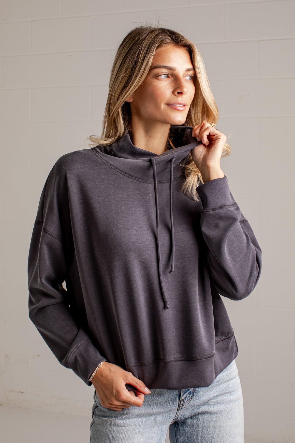 A woman wearing a dark charcoal gray oversized pullover hoodie with a high folded collar and drawstrings, paired with light blue jeans, standing against a light gray brick wall.