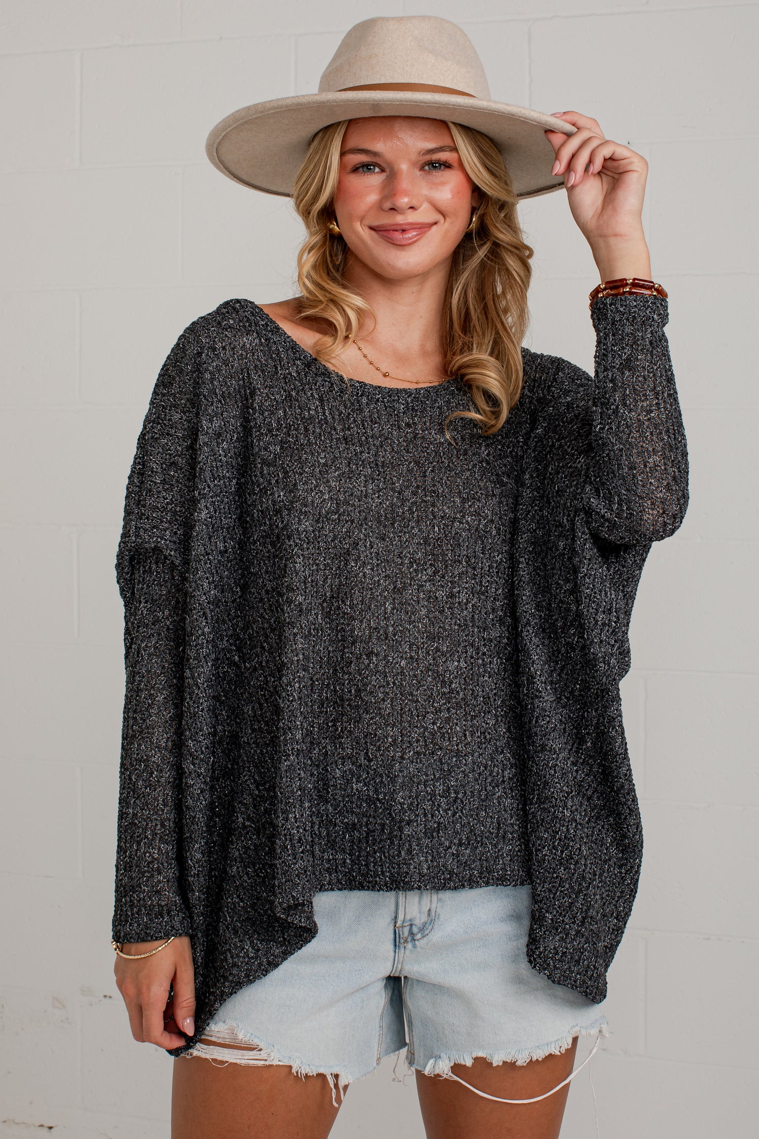 A woman wearing a dark gray heathered knit long-sleeve top, light-wash denim shorts, and a beige wide-brim hat, posing against a light gray tiled wall.