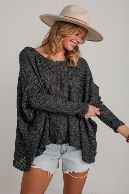 A woman wearing a dark gray heathered knit sweater with a wide boat neckline and long sleeves, paired with light-wash denim shorts and a beige wide-brim hat, posing against a white brick wall.