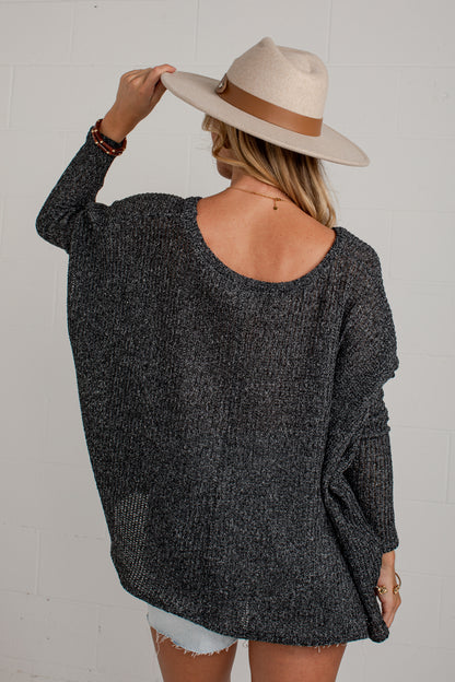 A woman shown from behind wearing a dark gray marled knit sweater, a beige wide-brim hat with a brown band, and light denim shorts against a white brick wall.