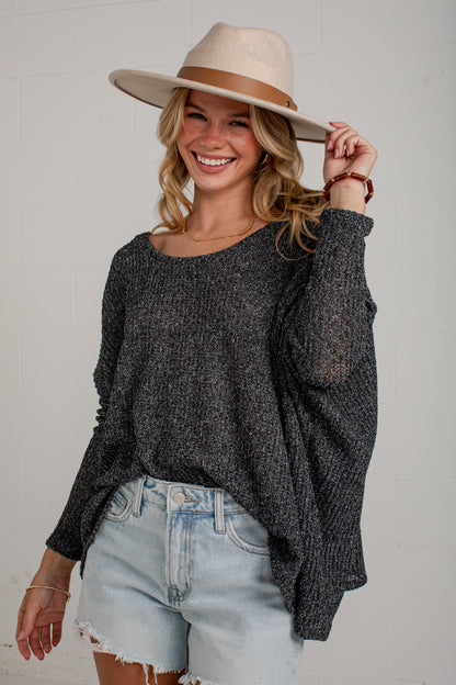 A smiling blonde woman wearing a dark charcoal gray knit sweater, light-wash denim shorts, and a beige wide-brim hat, posing against a white brick wall.