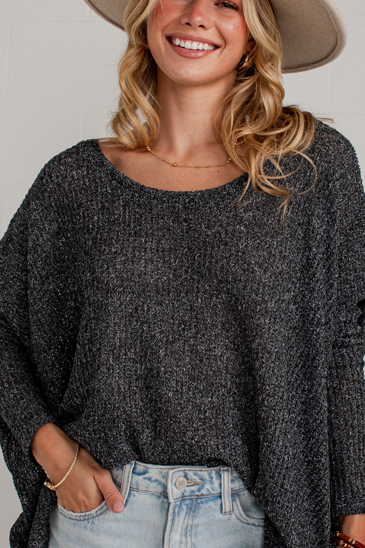 A woman wearing a dark gray marled knit long-sleeve top and light-wash jeans, with a beige wide-brim hat partially visible. The photo is cropped from the hat brim down to the hips, showing the texture of the knit top and the fit of the jeans against a light neutral background.