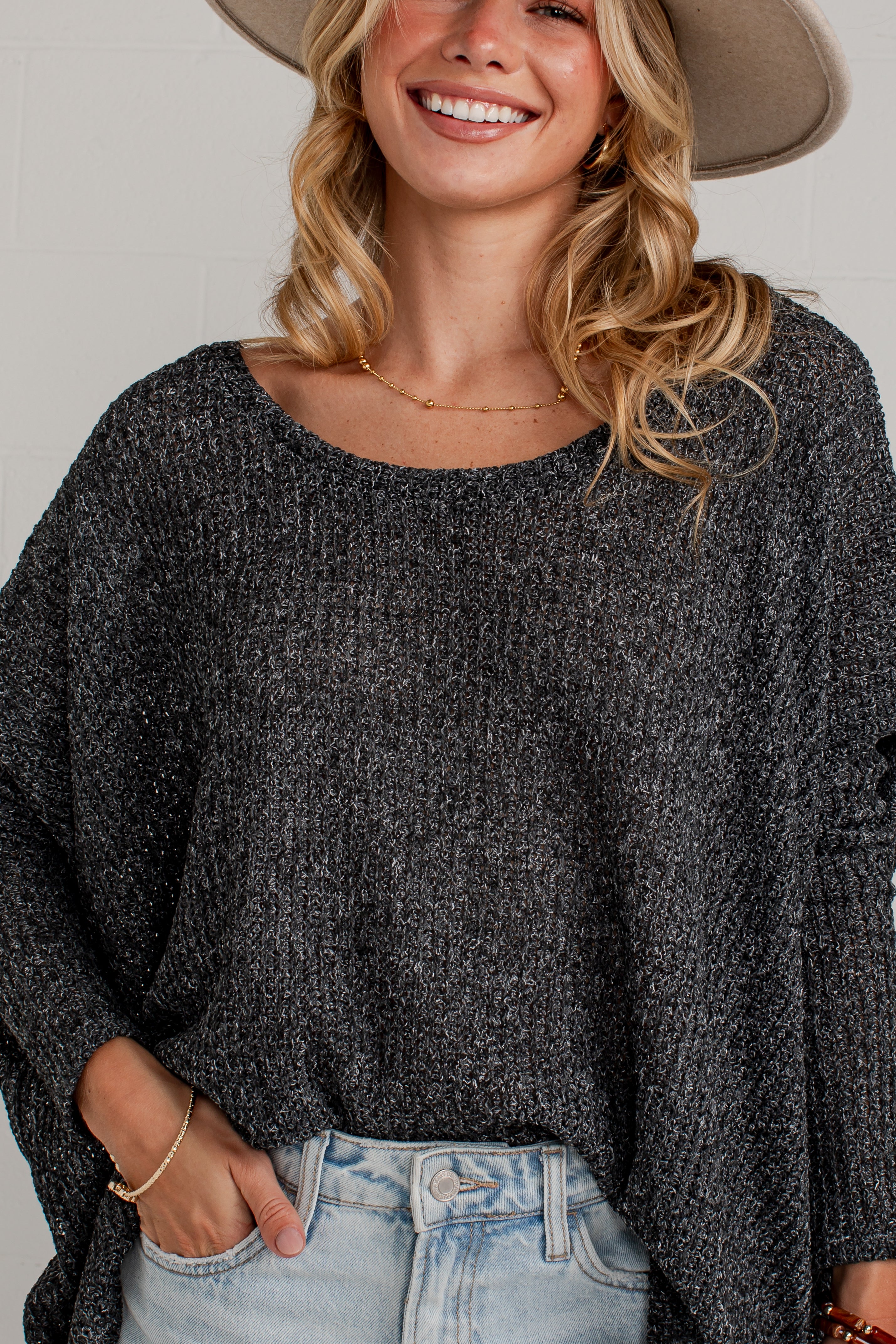 A woman wearing a dark gray marled knit long-sleeve top and light-wash jeans, with a beige wide-brim hat partially visible. The photo is cropped from the hat brim down to the hips, showing the texture of the knit top and the fit of the jeans against a light neutral background.