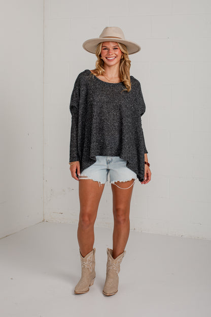 A woman modeling a dark gray heathered knit long-sleeve top, styled with light-wash denim shorts, beige cowboy boots, and a beige wide-brim hat, standing against a white studio wall.