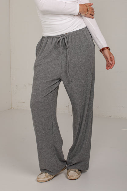 A person wearing loose, wide-leg gray drawstring sweatpants with a white long-sleeve top and beige shoes, standing in a plain indoor studio setting.