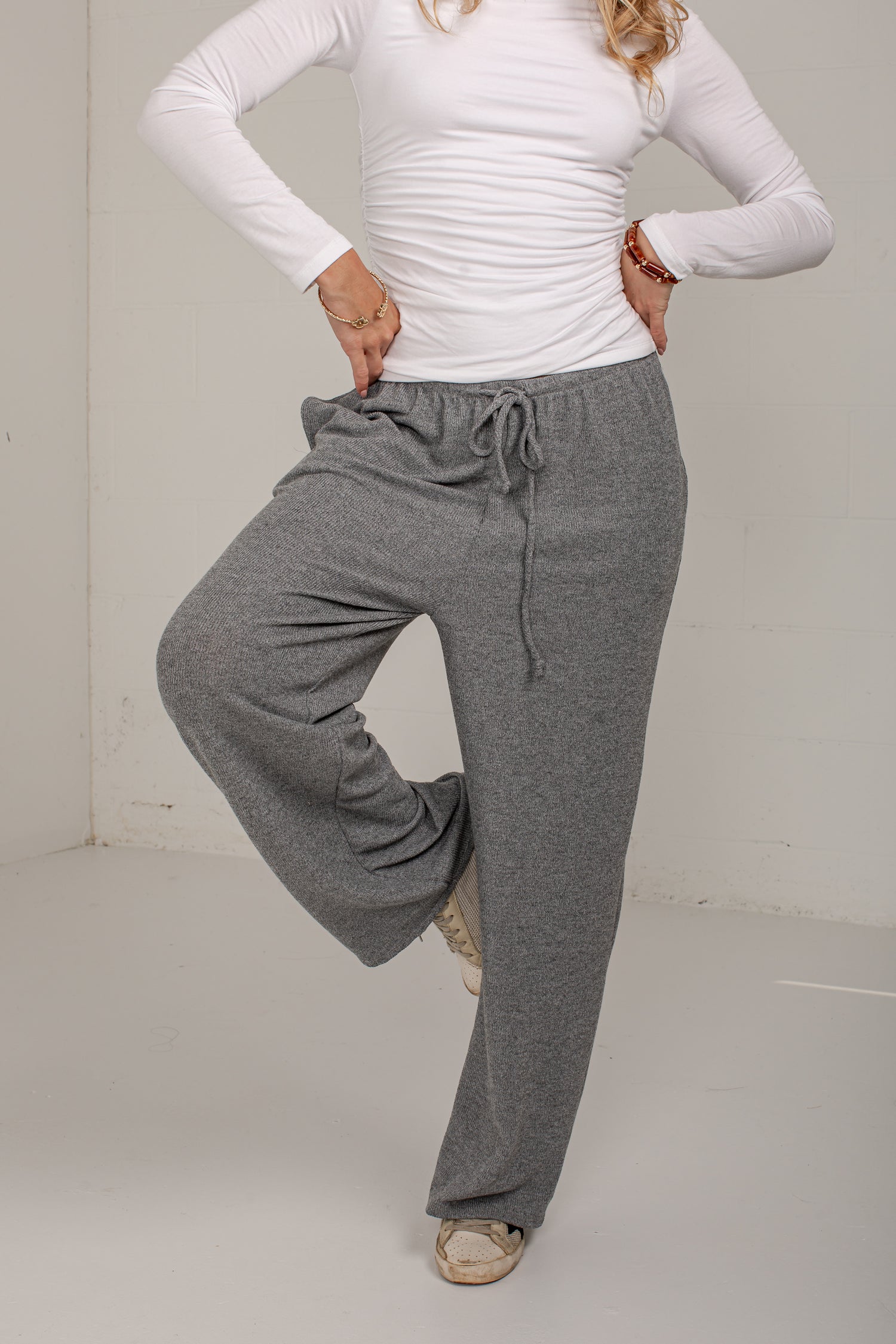 A woman modeling a pair of heather gray wide-leg lounge pants with a drawstring waist, paired with a white long-sleeve top and beige shoes.