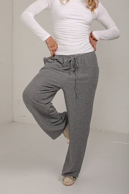 A woman modeling a pair of heather gray wide-leg lounge pants with a drawstring waist, paired with a white long-sleeve top and beige shoes.
