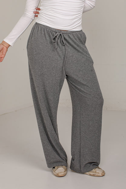 A person wearing loose, wide-leg gray drawstring sweatpants with a white long-sleeve top and beige shoes, photographed in a plain indoor studio setting.