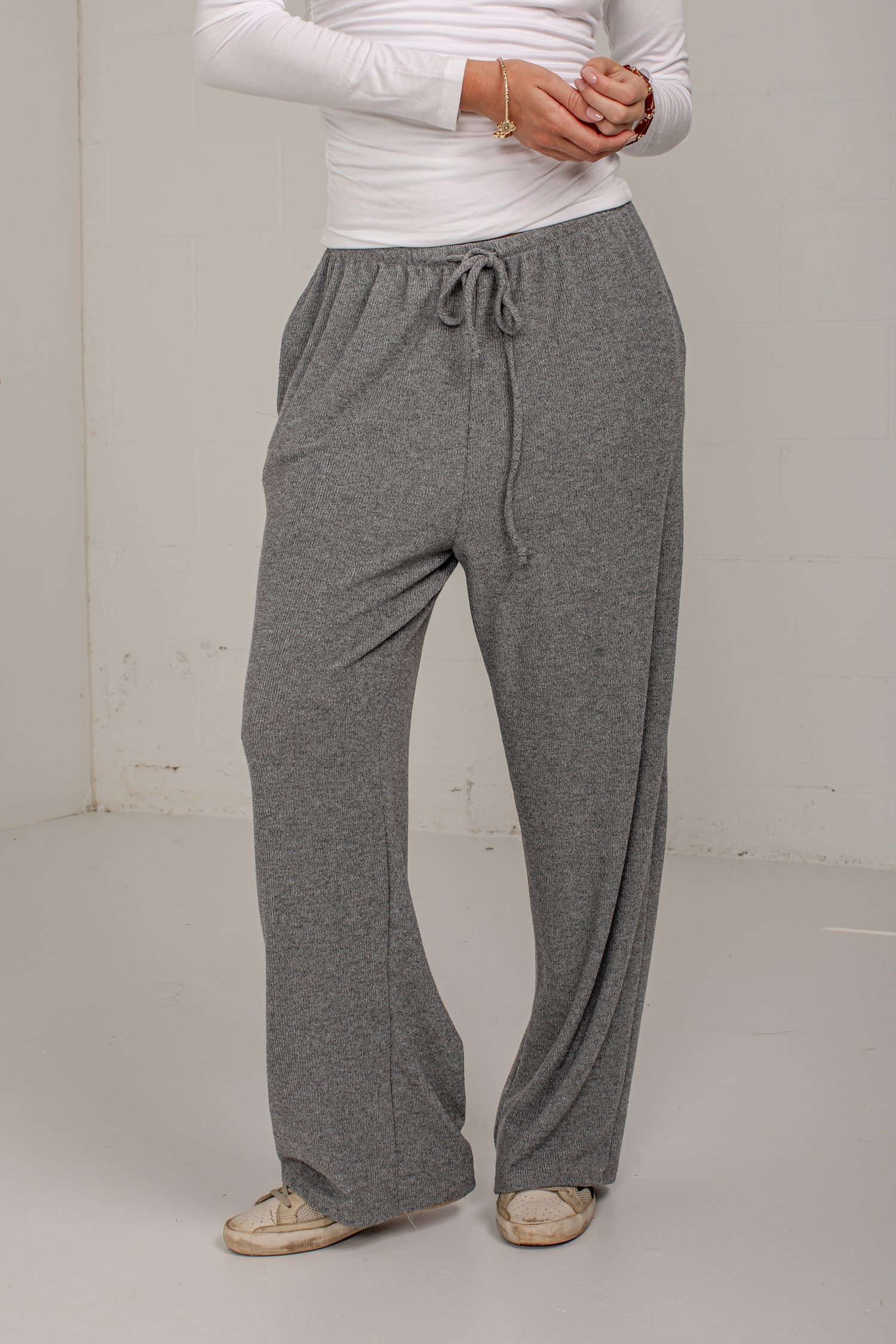 A person wearing heather gray drawstring lounge pants with a white long-sleeve top and beige shoes, standing against a plain off-white wall.