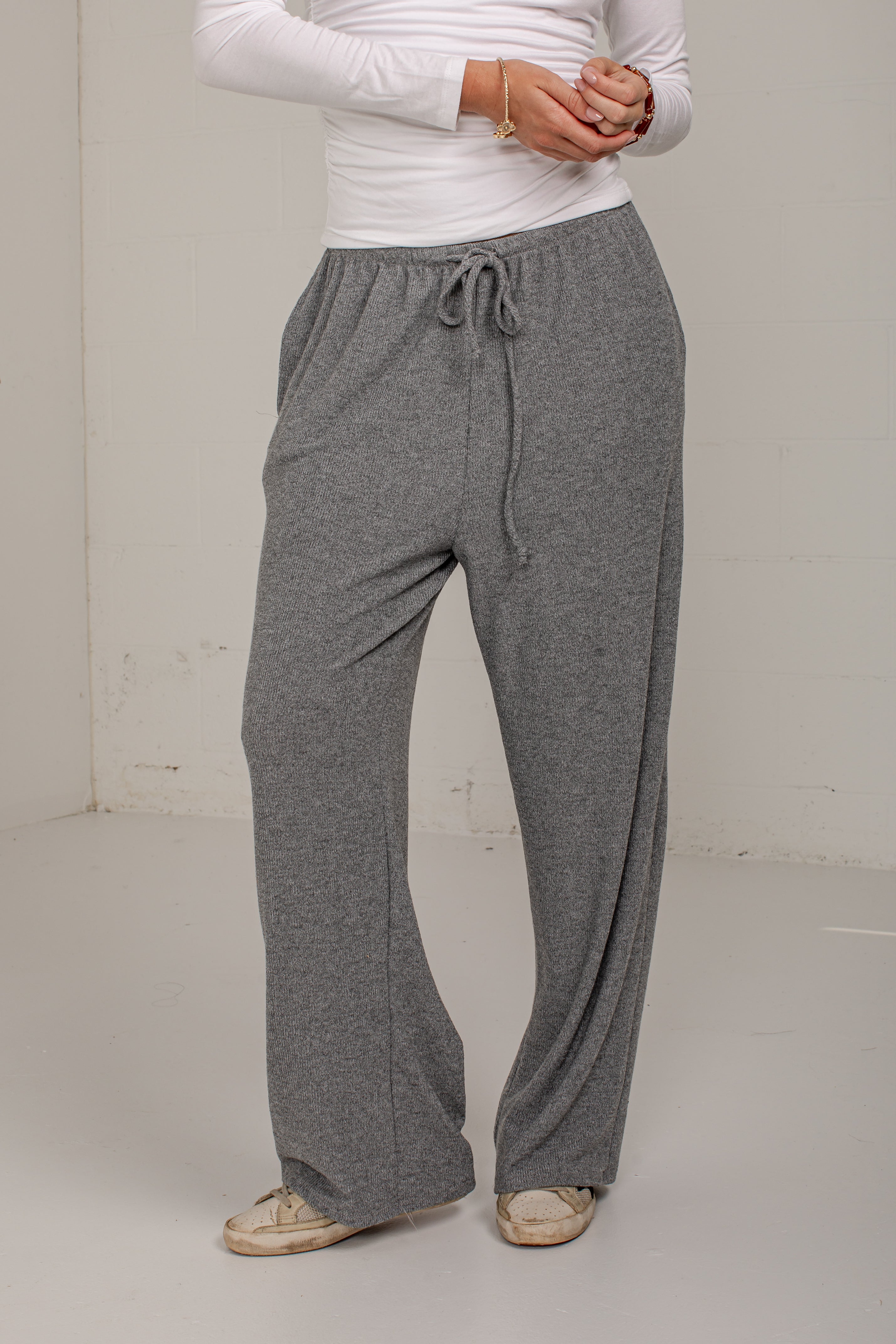 A person wearing heather gray drawstring lounge pants with a white long-sleeve top and beige shoes, standing against a plain off-white wall.