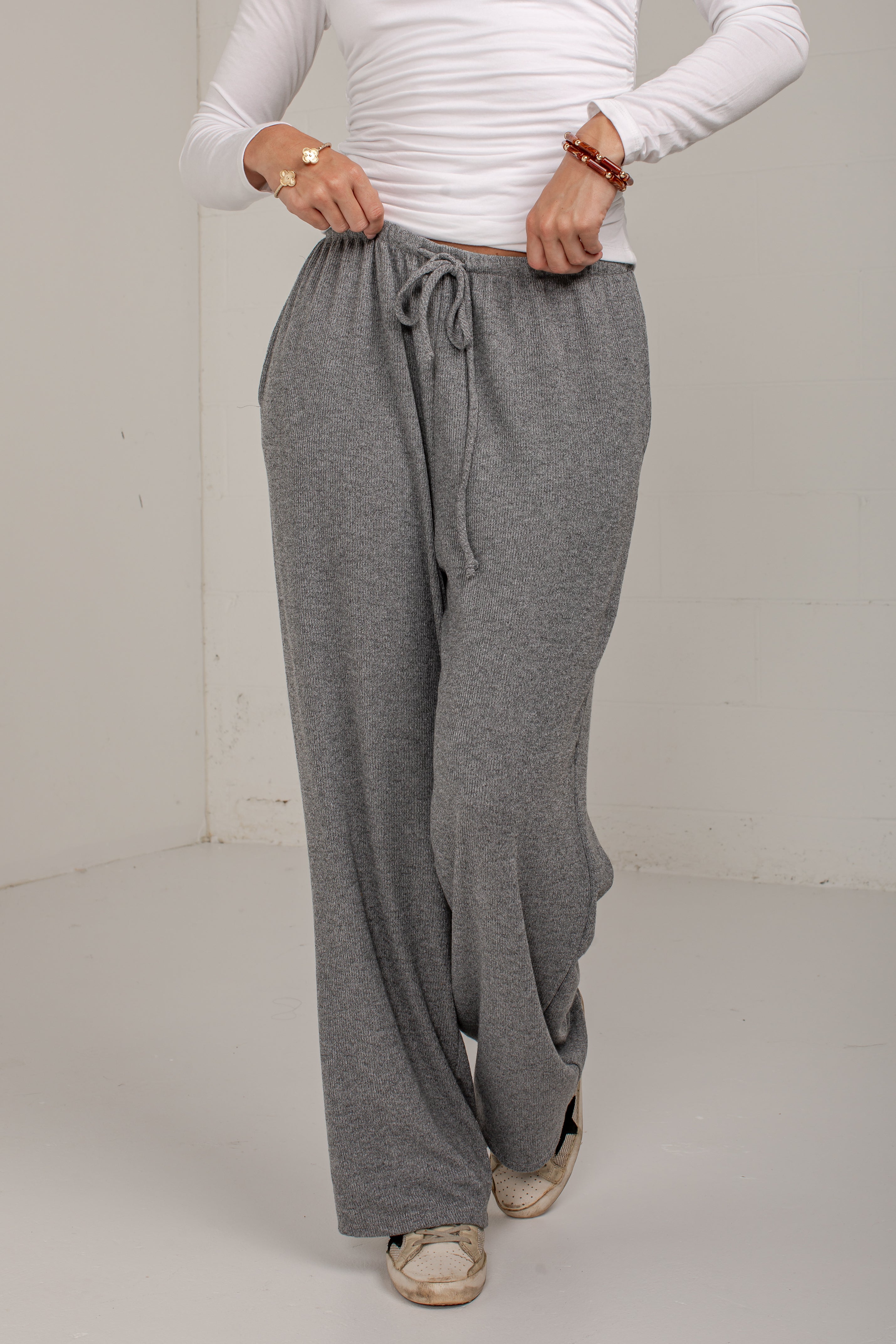 A person wearing loose, wide-leg gray drawstring sweatpants with a white long-sleeve top and beige shoes, standing against a plain off-white wall.