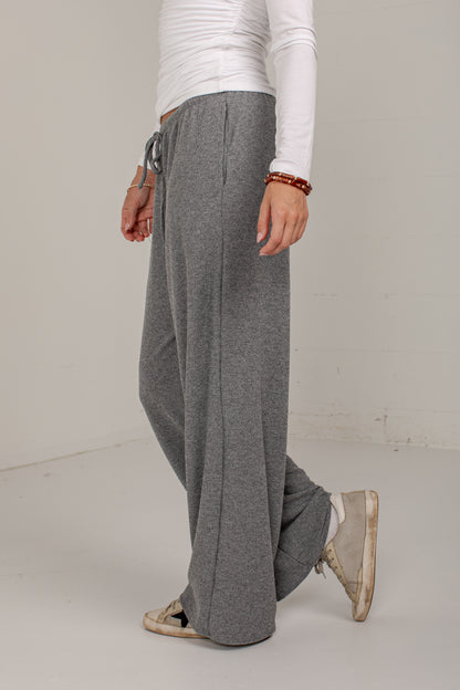 A person wearing loose, wide-leg gray sweatpants with a drawstring waist, paired with a white long-sleeve top and beige sneakers, standing against a plain off-white studio wall.
