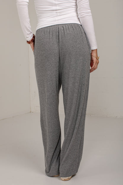 A person shown from the back wearing loose, wide-leg gray knit pants and a white long-sleeve top in a plain indoor setting.