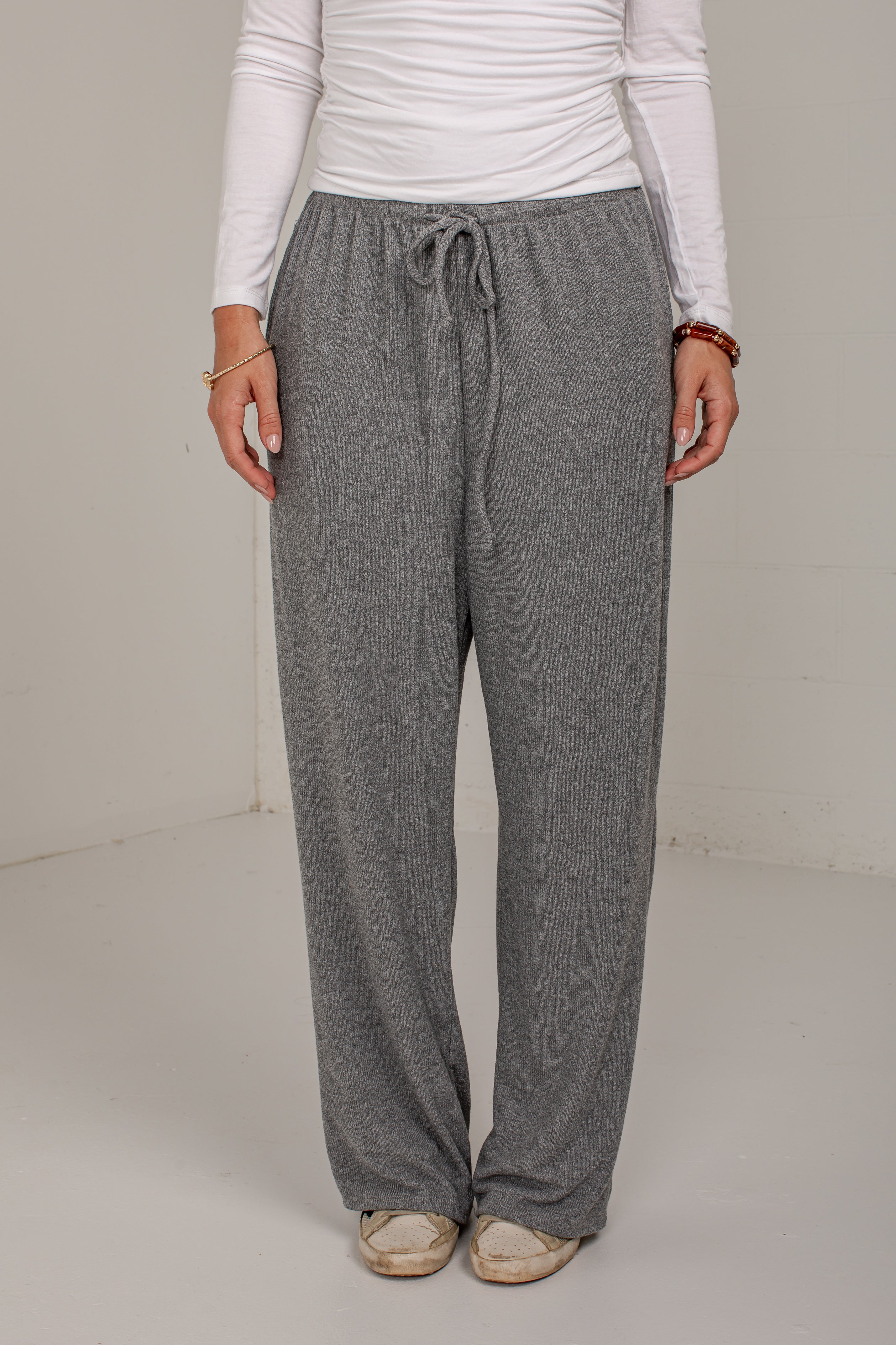 A person wearing heather gray drawstring lounge pants with a white long-sleeve top and beige shoes, standing against a plain light gray wall.