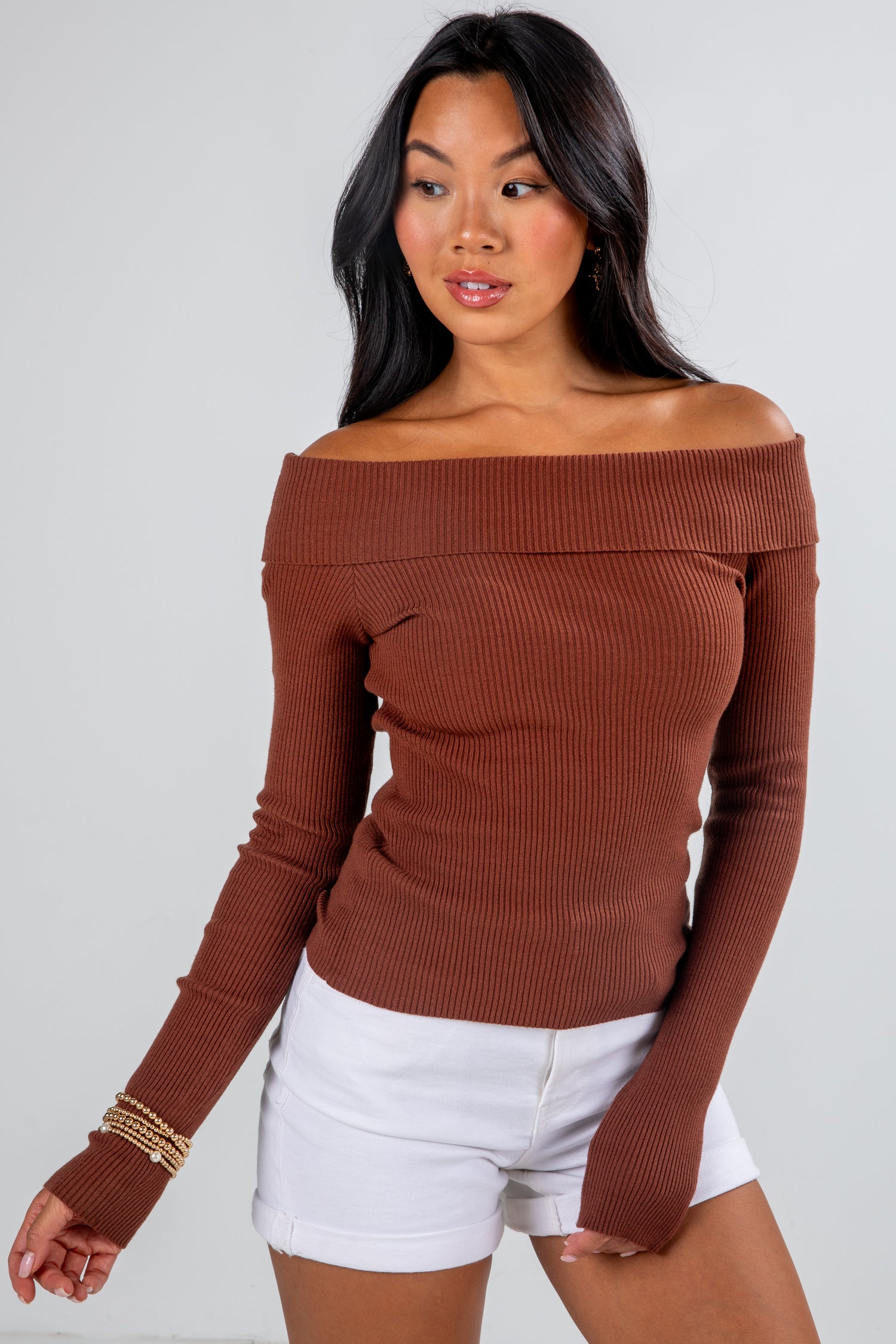 Woman wearing a brown off-shoulder sweater and white shorts on a plain background
