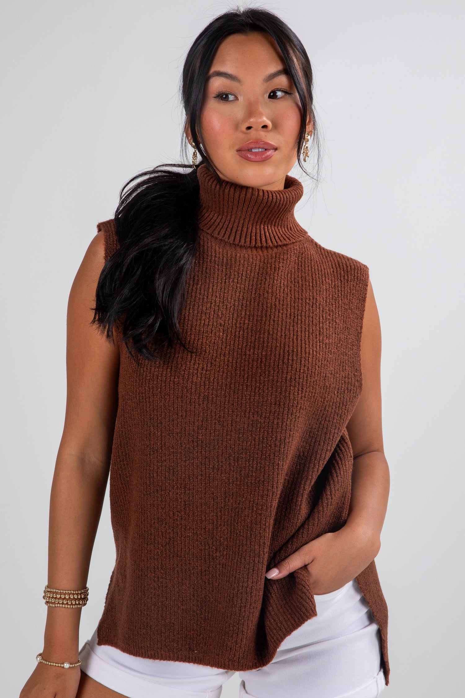 Woman wearing a brown sleeveless sweater over a white top against a plain background