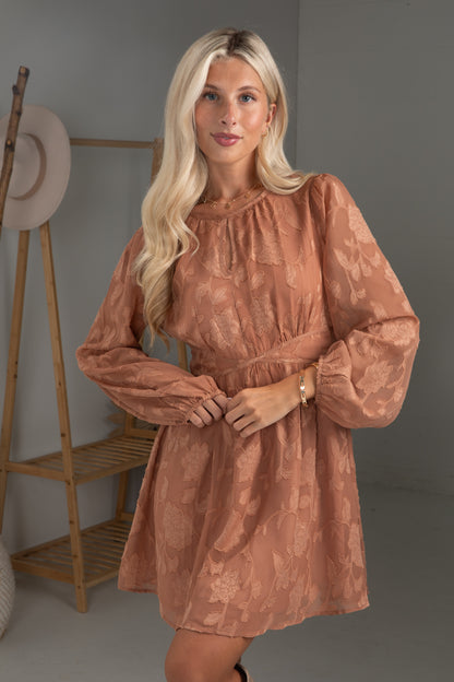 Woman wearing a brown dress standing in a room with a wooden shelf and hat.