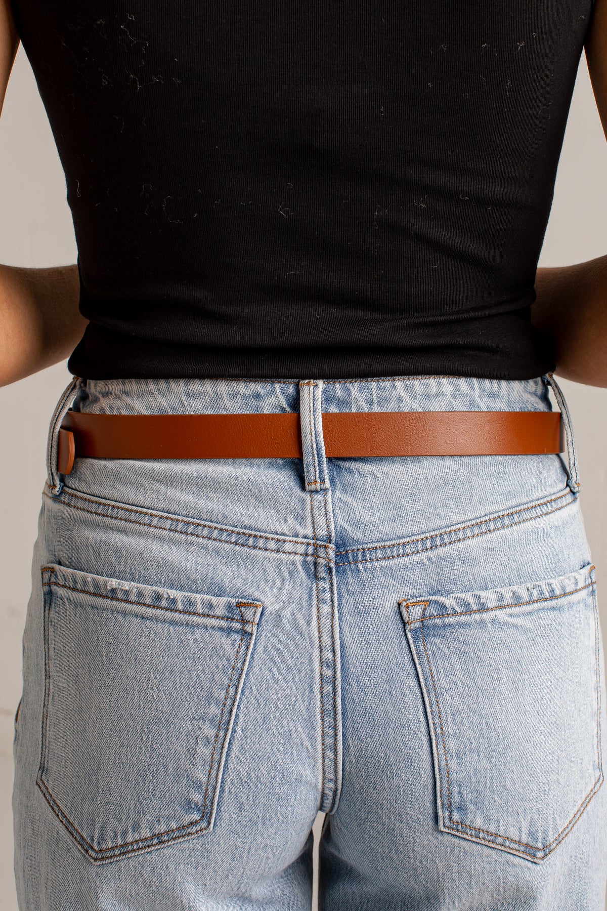 A close-up back view of a person wearing a slim brown leather belt with a small gold-tone accent on the left side, paired with light-wash blue jeans and a black ribbed top.
