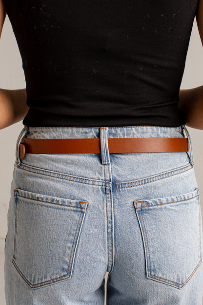 A close-up back view of a person wearing a slim brown leather belt with a small gold-tone accent on the left side, paired with light-wash blue jeans and a black ribbed top.