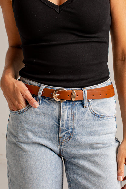A close-up of a person wearing a slim brown leather belt with a polished gold-tone oval buckle, styled with light-wash blue jeans and a black ribbed tank top.
