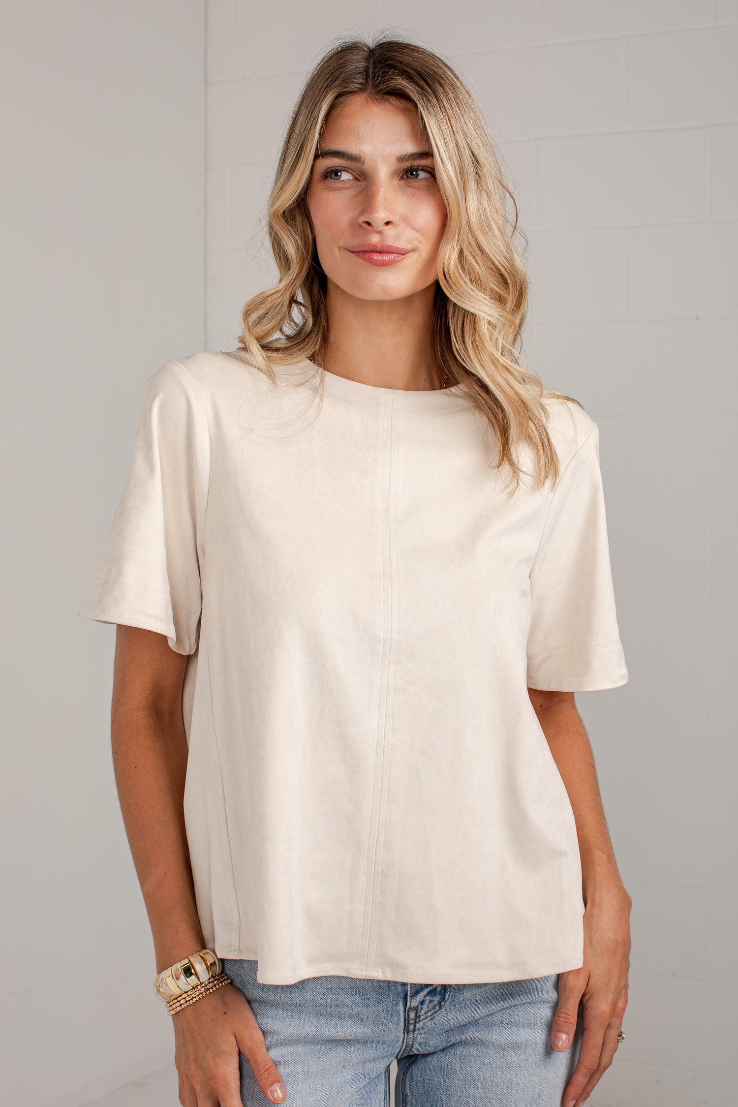 A woman wearing a cream-colored short-sleeve top and light blue jeans, standing against a light gray wall.