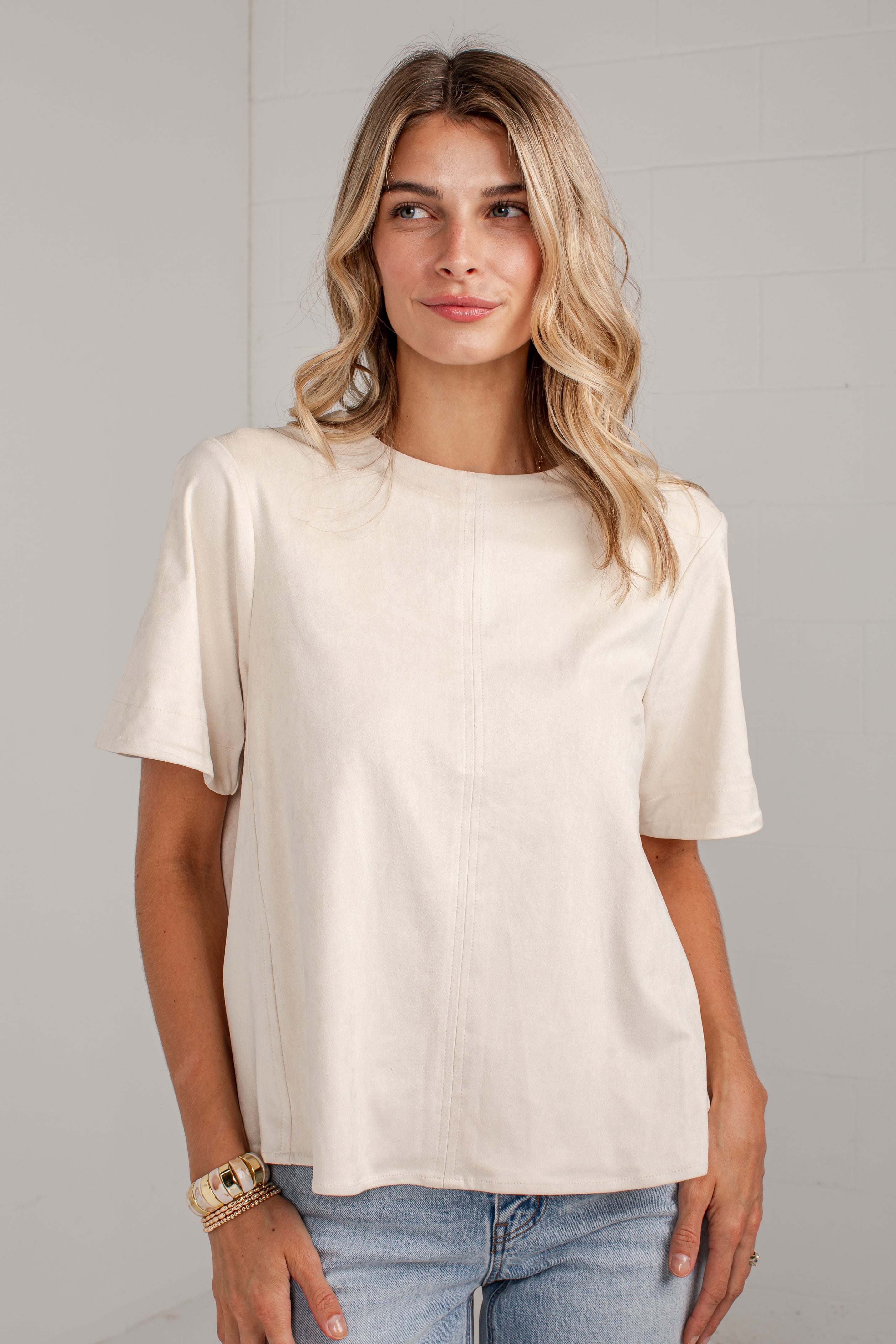 A woman wearing a cream-colored short-sleeve top and light blue jeans, standing against a light gray wall.