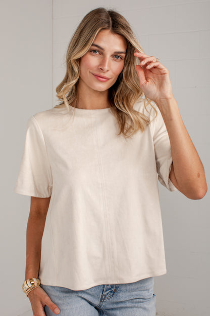 A woman wearing a short-sleeve cream-colored top and light blue jeans, standing against a light gray paneled wall.