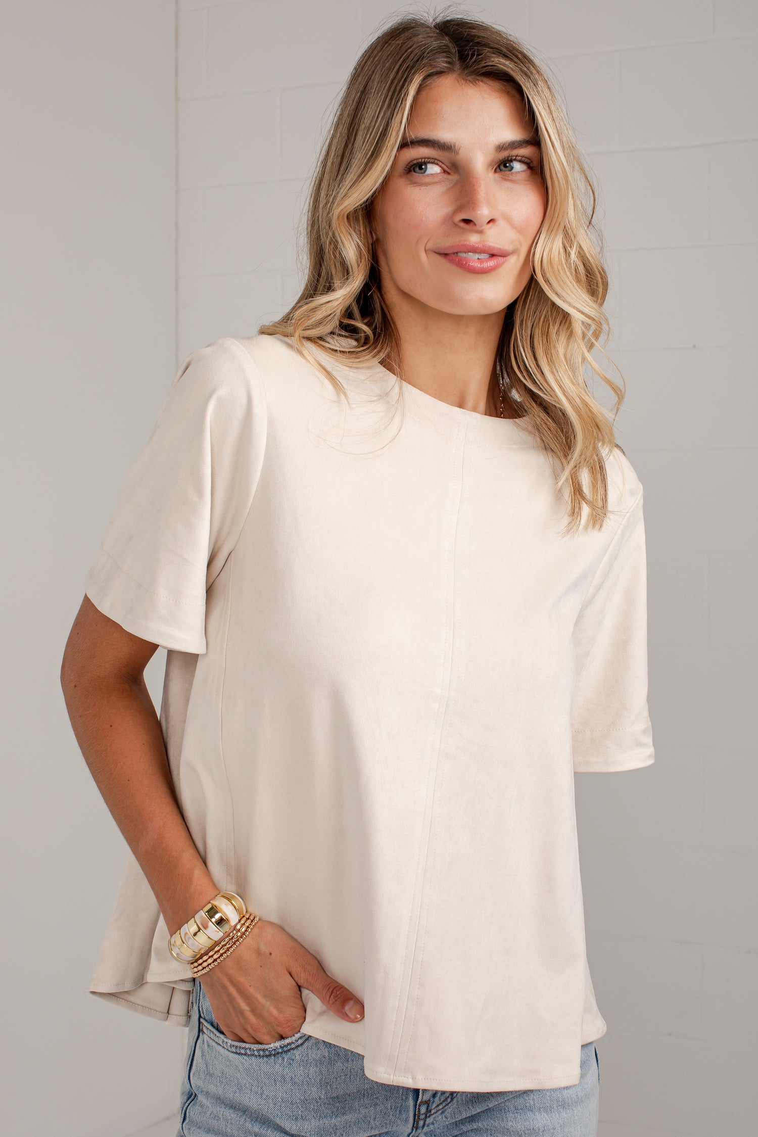 A woman wearing a short-sleeve cream-colored top and light blue jeans, standing against a light gray paneled wall.