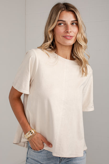 A woman wearing a short-sleeve cream-colored top and light blue jeans, standing against a light gray paneled wall.