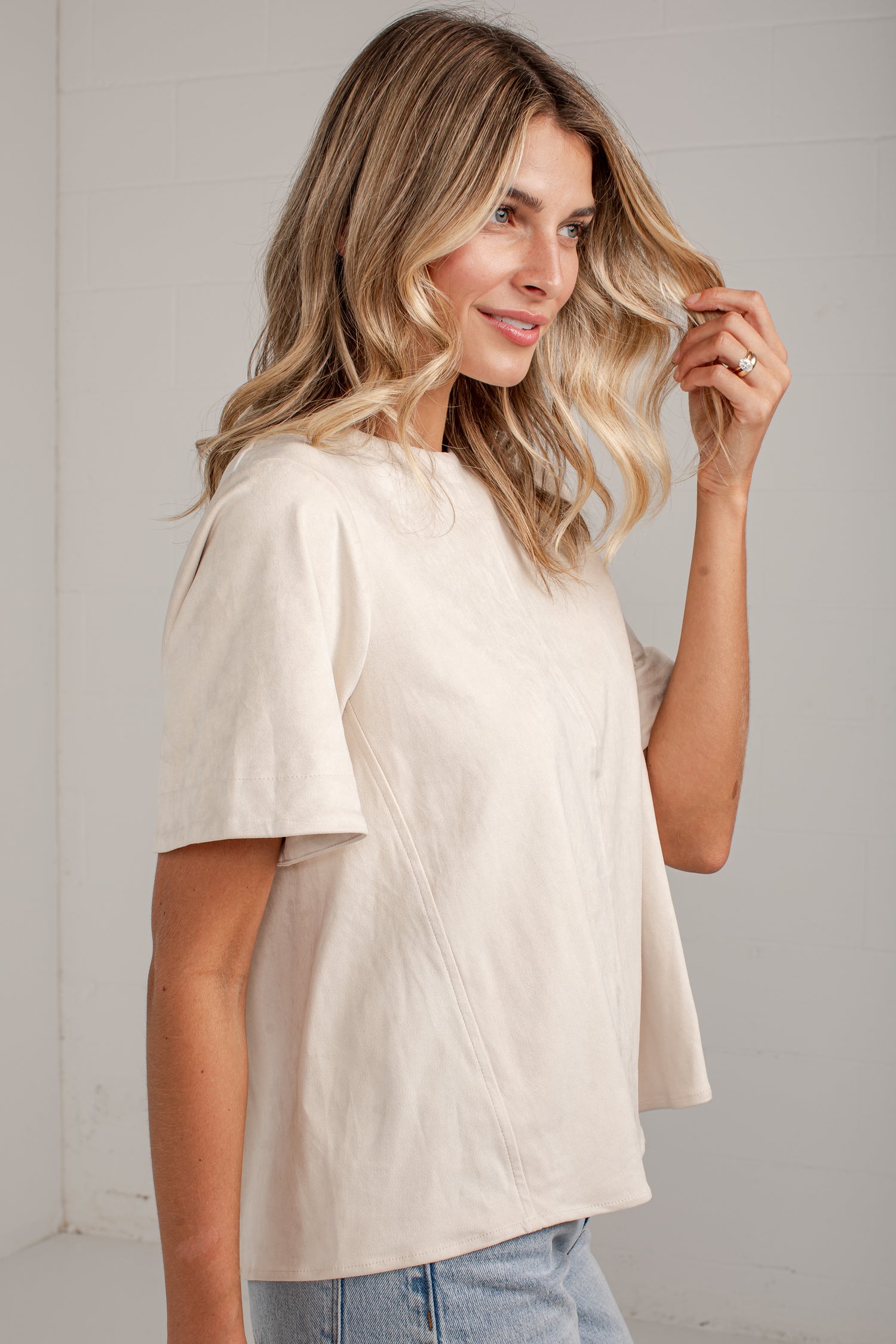 A woman wearing a short-sleeve cream-colored top with diagonal seam details, paired with light blue jeans, standing against a white paneled wall.