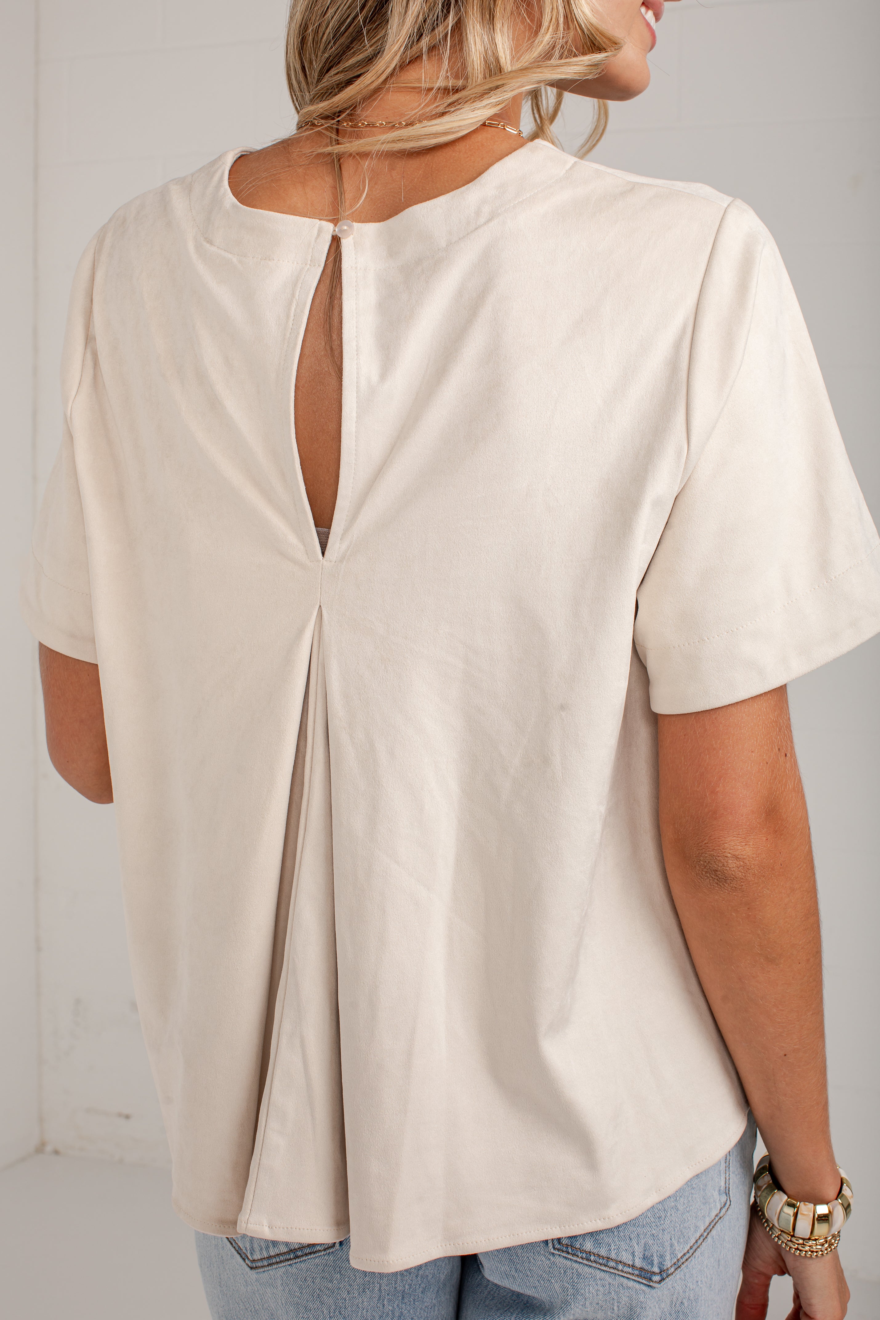 A woman wearing a cream-colored short-sleeve top with a back keyhole cutout, shown from the back against a plain light background.