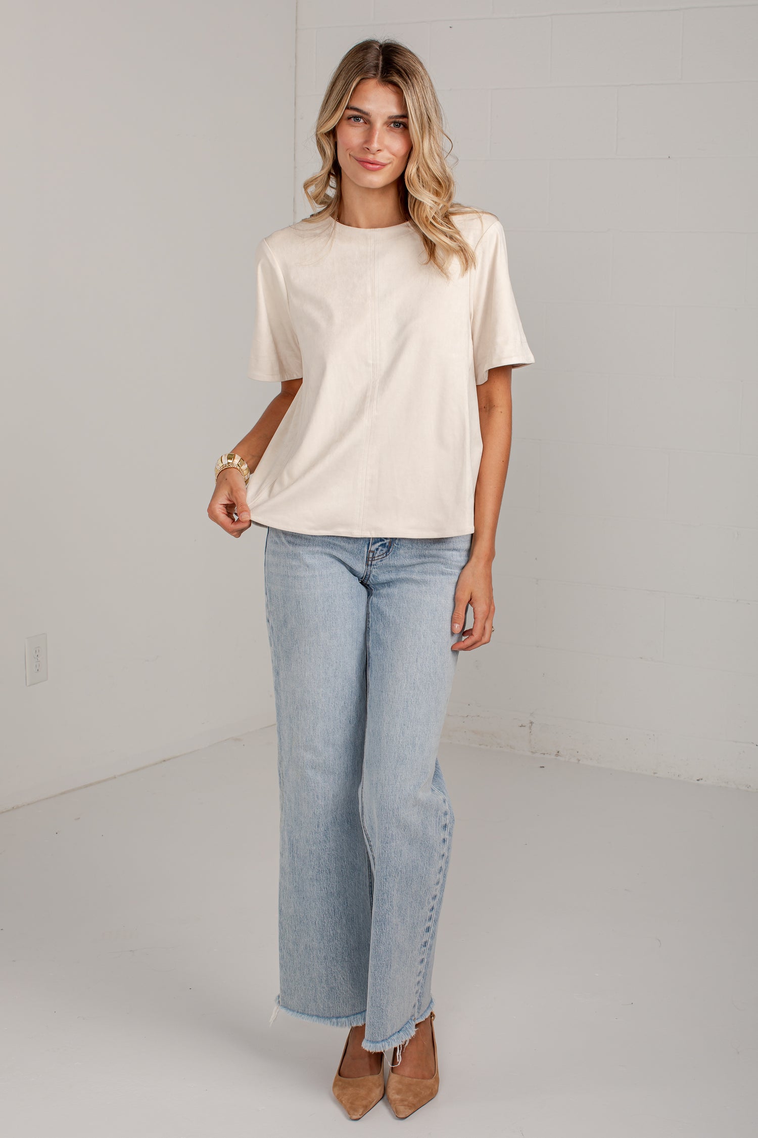 A woman modeling a cream-colored short-sleeve crewneck t-shirt and light-wash wide-leg jeans with side button details, paired with brown pointed-toe heels, in a bright white studio setting.