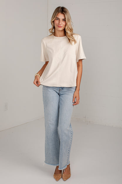 A woman modeling a cream-colored short-sleeve crewneck t-shirt and light-wash wide-leg jeans with side button details, paired with brown pointed-toe heels, in a bright white studio setting.
