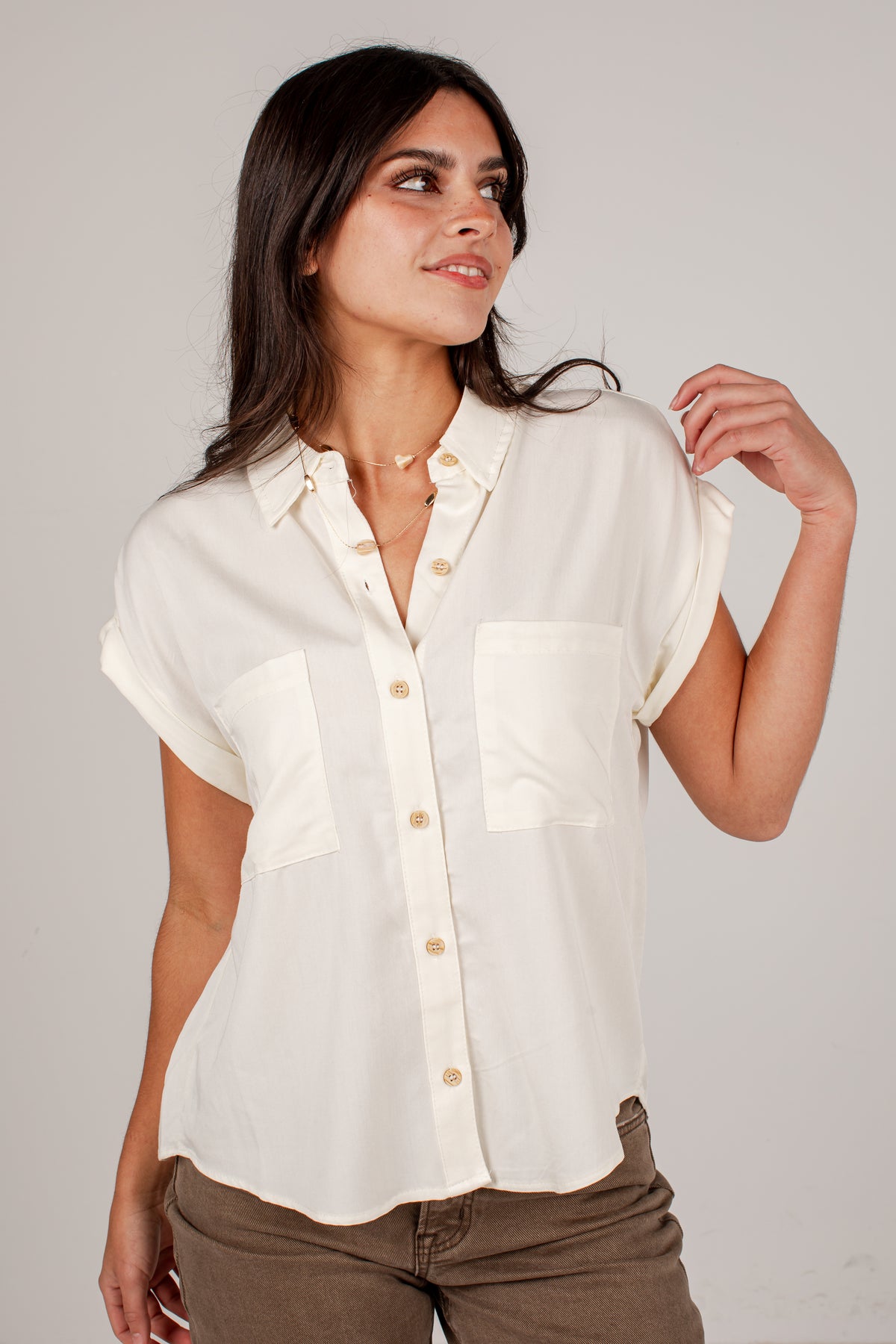 A woman wearing a short-sleeve cream-colored button-up shirt with two chest pockets, paired with brown pants, photographed against a neutral light gray studio background.
