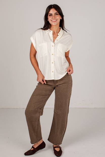 A woman modeling a short-sleeve cream button-up shirt and brown wide-leg pants against a light gray studio background.