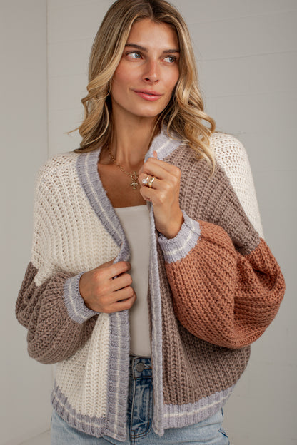 A woman wearing a chunky knit cardigan with a color-blocked design in cream, taupe, and terracotta, styled over a light gray top and blue jeans.