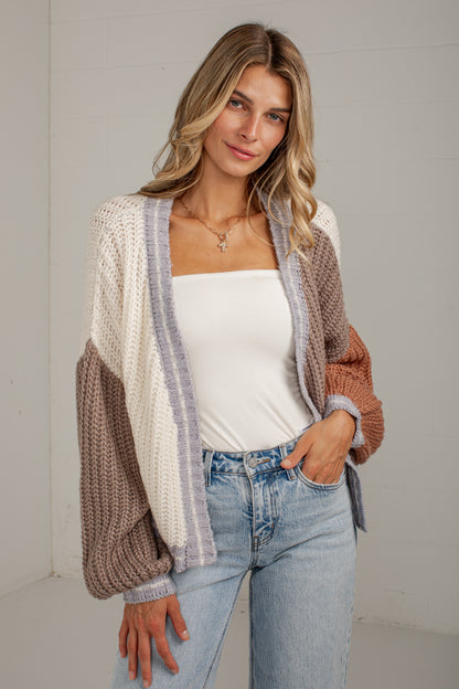 A woman modeling a chunky knit color-block cardigan with cream, taupe, and rust-brown panels, worn over a white top and light-wash blue jeans against a light gray wall.