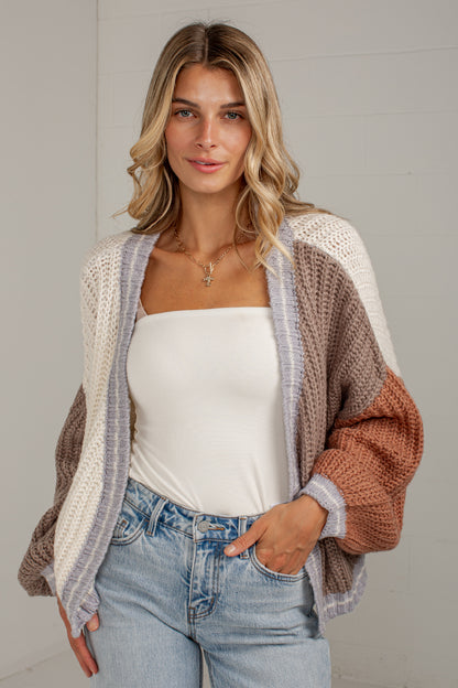 A woman wearing a chunky knit color-block cardigan with beige, brown, and white panels, styled over a white tube top and light blue jeans.