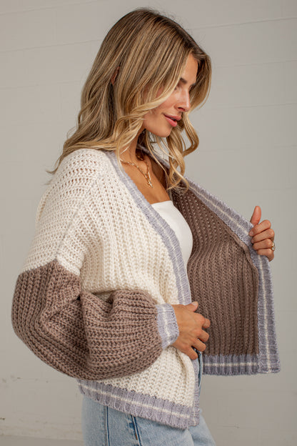 A woman wearing a chunky knit color-block cardigan with cream, taupe, and light gray stripes, holding the front open to show the interior lining.