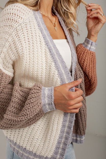 A close-up of a woman wearing a chunky knit cardigan with a cream body, taupe sleeves, and gray plaid trim, layered over a white top.